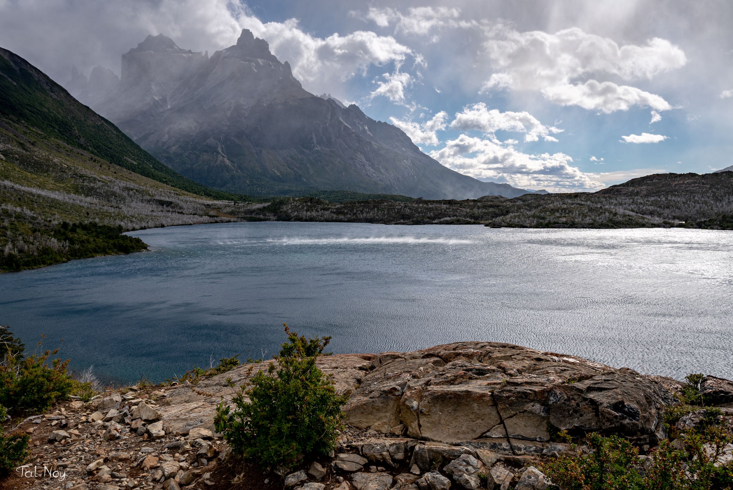 A lake surrounded by rugged terrain with the jagged peaks of Torres del Paine shrouded in clouds above
